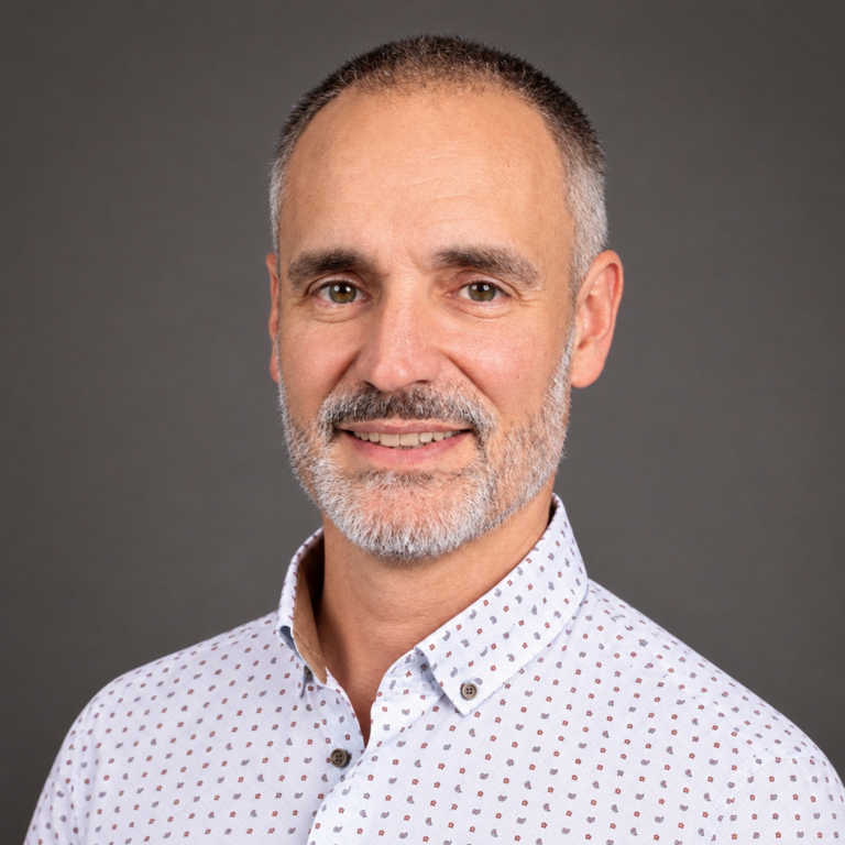 Homme souriant avec une chemise à motifs, cheveux gris et barbe courte.