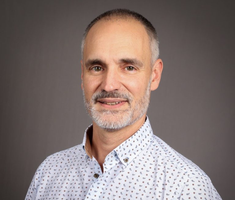 Homme souriant avec une chemise à motifs, cheveux gris et barbe courte.