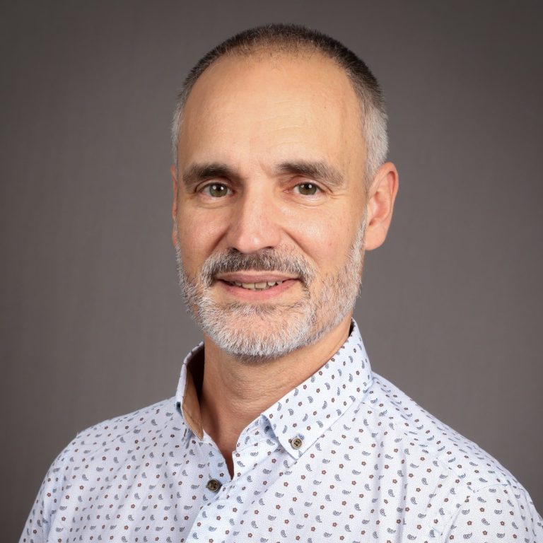 Homme souriant avec une chemise à motifs, cheveux gris et barbe courte.