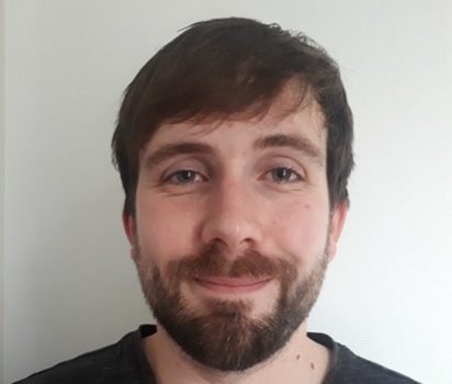 Homme souriant avec une barbe, vêtu d'un t-shirt noir, devant un fond blanc.